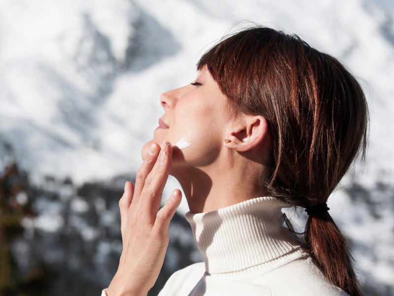 Une femme avec un pull à col roulé applique de la crème sur son visage et en arrière-plan, il y a de la neige sur les montagnes