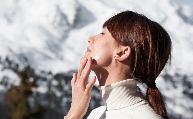 Une femme avec un pull à col roulé applique de la crème sur son visage et en arrière-plan, il y a de la neige sur les montagnes